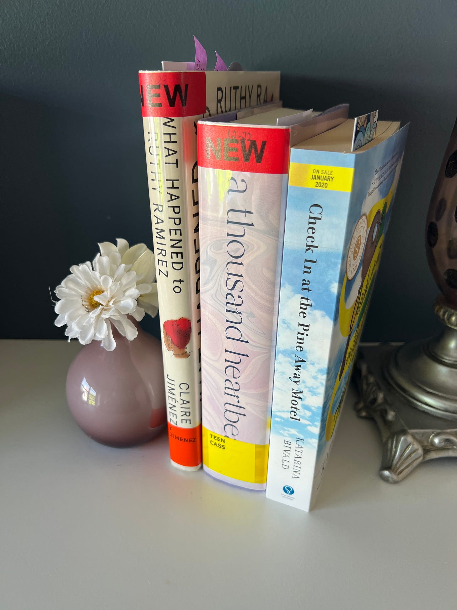The books What Happened to Ruthy Ramirez, A Thousand Heartbeats, and Welcome to the Pine Away Motel and Cabins, standing upright next to a tiny purple vase with a white flower. 