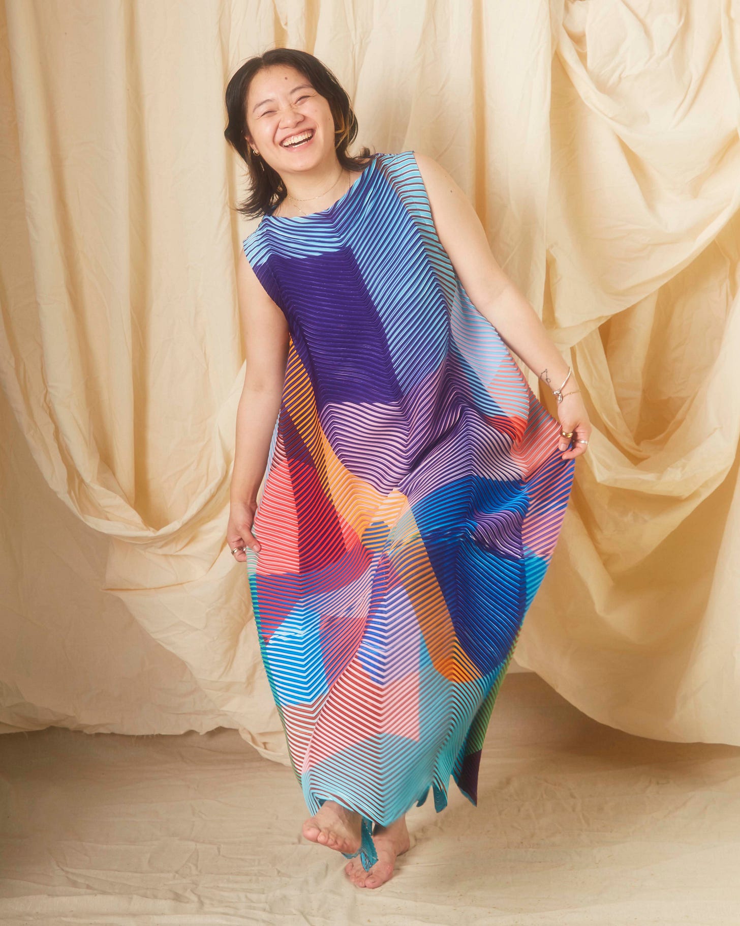Ka Yee Chan stands in front of a beige background wearing a multi-color dress where she holds up both sides and laughs towards the camera. Ka Yee Chan stands in front of a beige background wearing a multi-color dress where she holds up both sides and laughs towards the camera.