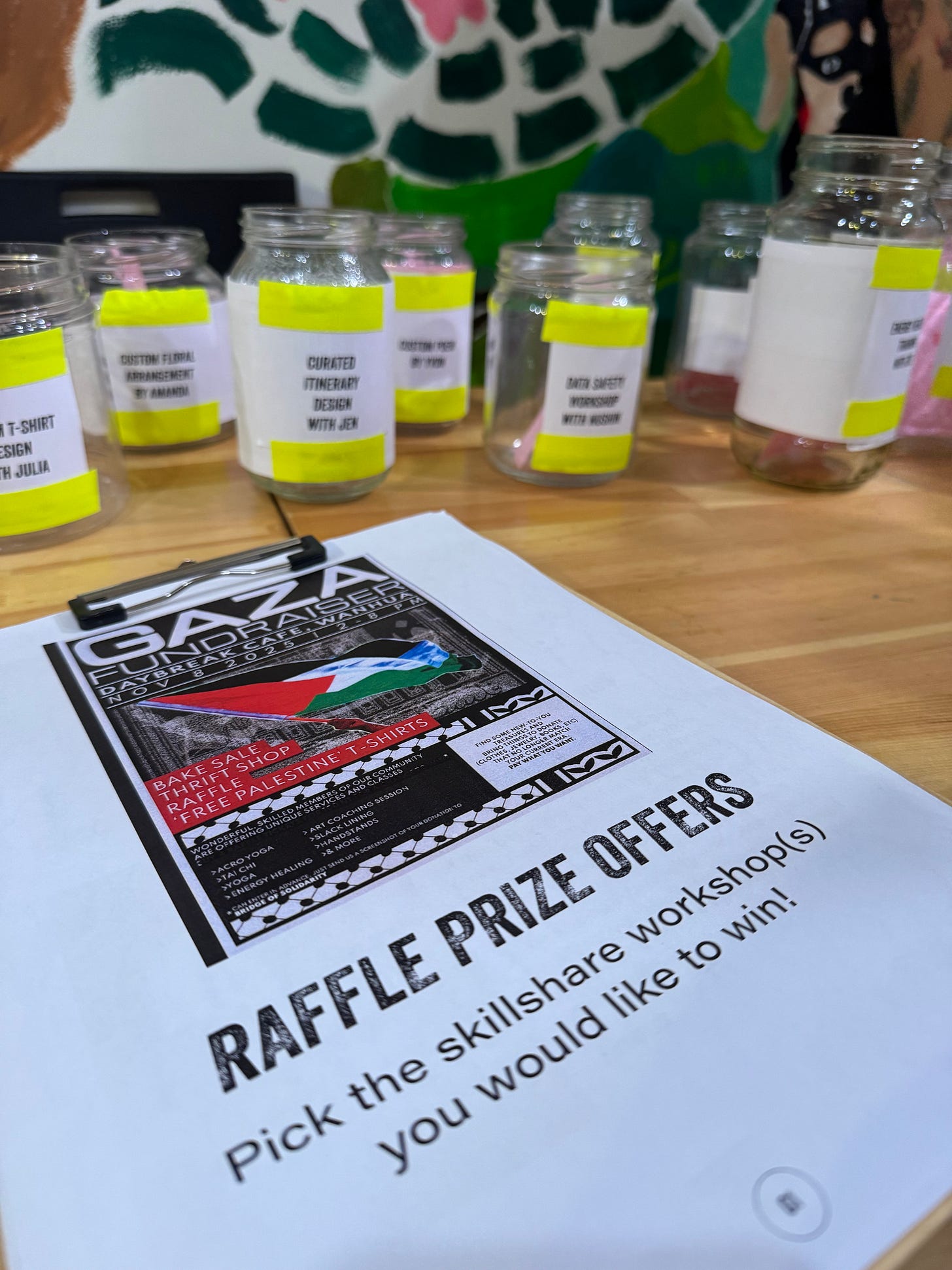 Vertical photo of a Gaza fundraiser poster reading Raffle Prize offers, taped before a table of jars for donations to different community prizes. 