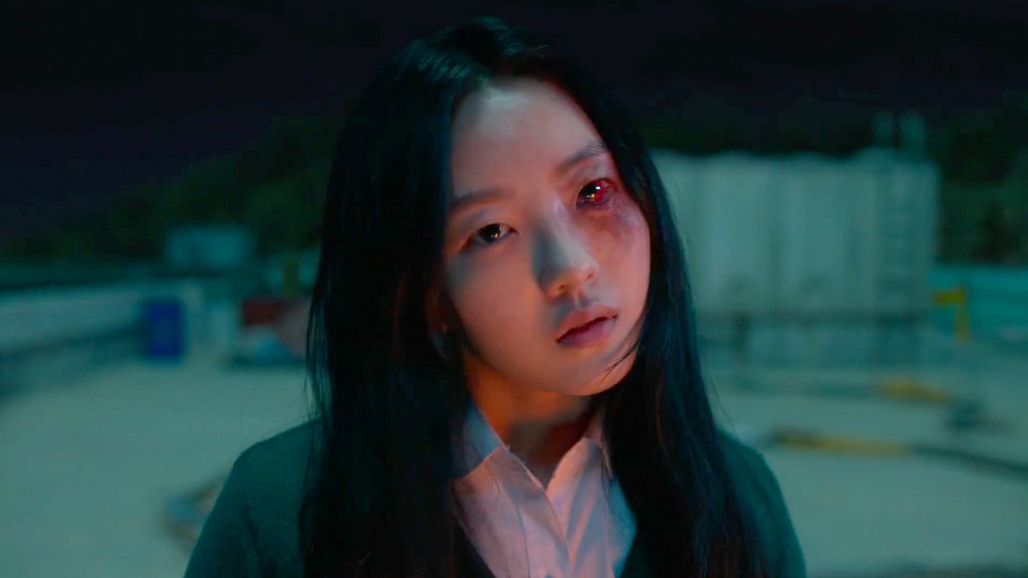 A young girl with a red, bloody, and irritated eye looks at the camera, her head cocked to the side in a sinister way. She looks to be inside an empty parking lot at night, with many trees in the distance. Still from the show All of Us Are Dead