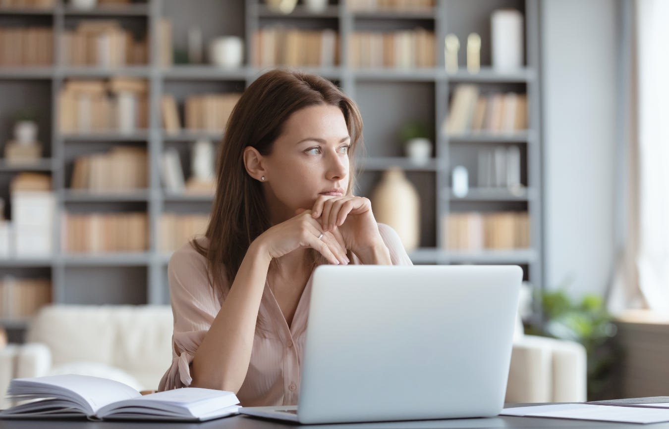 A woman at a desk looking off in the distance pensively, natural light, calm not chaotic. She has an open laptop and notebook in front of her, a visual of how much she has to do.