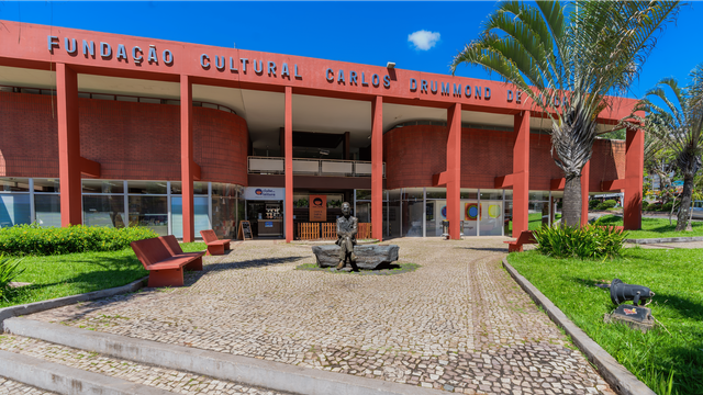 #descrevemospravocê  Fachada da Fundação Cultural Carlos Drummond de Andrade, um prédio público de dois andares com arquitetura moderna, localizado em Itabira, Minas Gerais. A construção é marcada por colunas verticais na cor vermelho-alaranjada e paredes curvas em tijolos aparentes. A parte inferior é composta por vitrines e portas de vidro, com diversos cartazes e placas indicando atividades culturais e exposições. Acima da entrada principal, há uma marquise sustentada pelas colunas. No topo do prédio, em letras maiúsculas e espaçadas, está escrito: “FUNDAÇÃO CULTURAL CARLOS DRUMMOND DE ANDRADE”.  Em frente ao edifício, no centro de uma pequena praça de piso em pedras portuguesas, há uma escultura de um homem sentado em um banco de pedra. A escultura representa o poeta Carlos Drummond de Andrade, com expressão serena, usando óculos e vestindo paletó. Ao redor, há bancos vermelhos de concreto e canteiros com grama bem cuidada. No lado direito da imagem, vê-se uma palmeira alta e luminárias de jardim. O céu está azul com algumas nuvens pequenas e esparsas, e o ambiente é iluminado pela luz do sol.