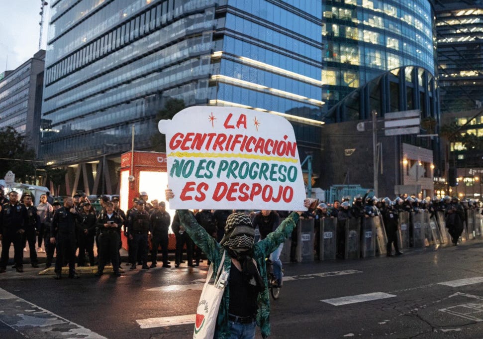 Una persona manifestante con il volto coperto da una kefiah mostra un cartello con scritto "Gentrificación no es progreso, es despojo" nel tipico stile dei rotulos messicani. Dietro di lei, polizia e grattacieli di Città del Messico.