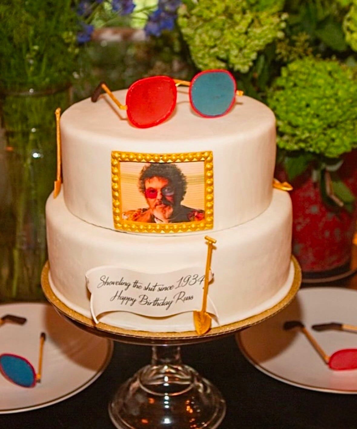 Russ Tamblyn's 90th birthday cake. A two-layer, circular white cake with gold shovels around the exterior. On the top of the cake is an edible version of Dr. Jacoby's red-and-blue lens eyeglasses. The front of the cake has a photo of Russ in character as Dr. Jacoby. Under the photo is an edible plaque that reads "Shoveling the shit since 1934. Happy birthday, Russ"
