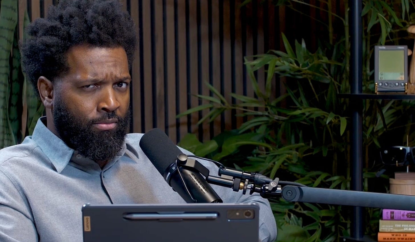 Baratunde Thurston in the Life With Machines studio, sitting behind a Lenovo tablet and microphone with a skeptical expression on his face. Plants and books visible in the background. Screenshot from the post-show conversation with AI co-producer BLAIR after the Sam Gregory interview. Baratunde Thurston in the Life With Machines studio, sitting behind a Lenovo tablet and microphone with a skeptical expression on his face. Plants and books visible in the background. Screenshot from the post-show conversation with AI co-producer BLAIR after the Sam Gregory interview.