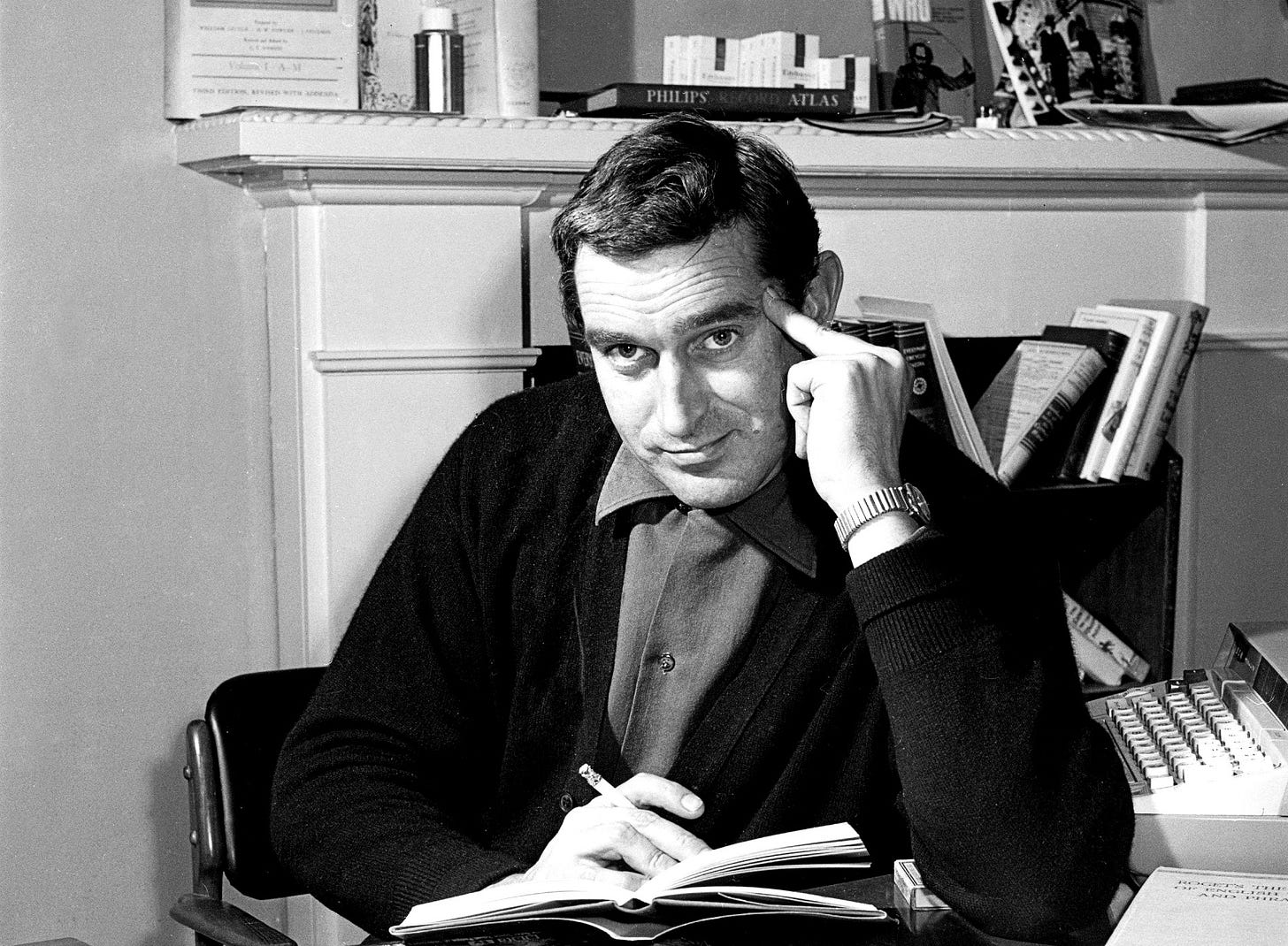 A youngish white man (Terry Nation) sitting in an office with typewriter and various books close to hand, and smoking a cigarette