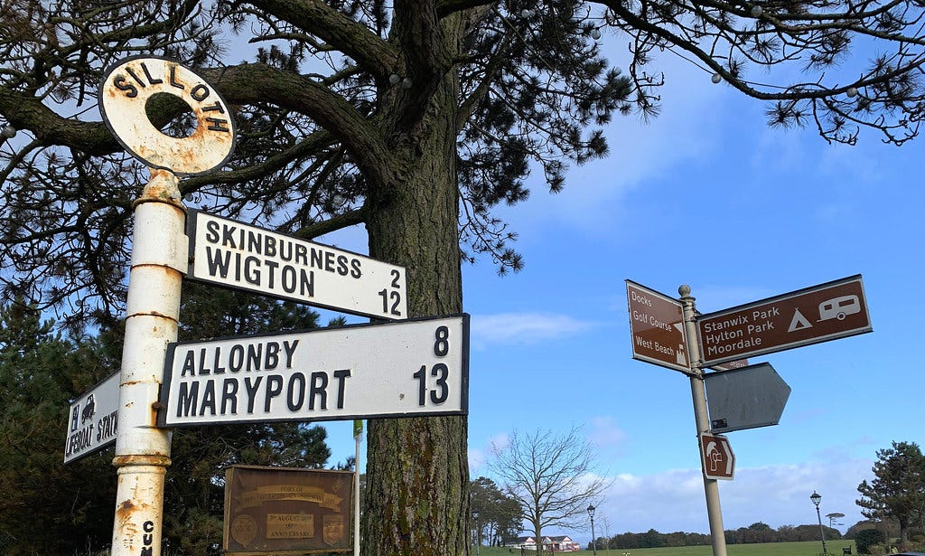 A 19th or 20th century fingerpost-style sign with "Silloth" written on the roundel, a brown tourist roadsign from the modern day is in the background along with trees and a village green A 19th or 20th century fingerpost-style sign with "Silloth" written on the roundel, a brown tourist roadsign from the modern day is in the background along with trees and a village green