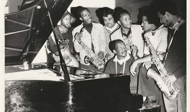 An old black and white photo of young to middle-aged African men, one seated at a paino and surrounded by the others, some holding musical instruments.