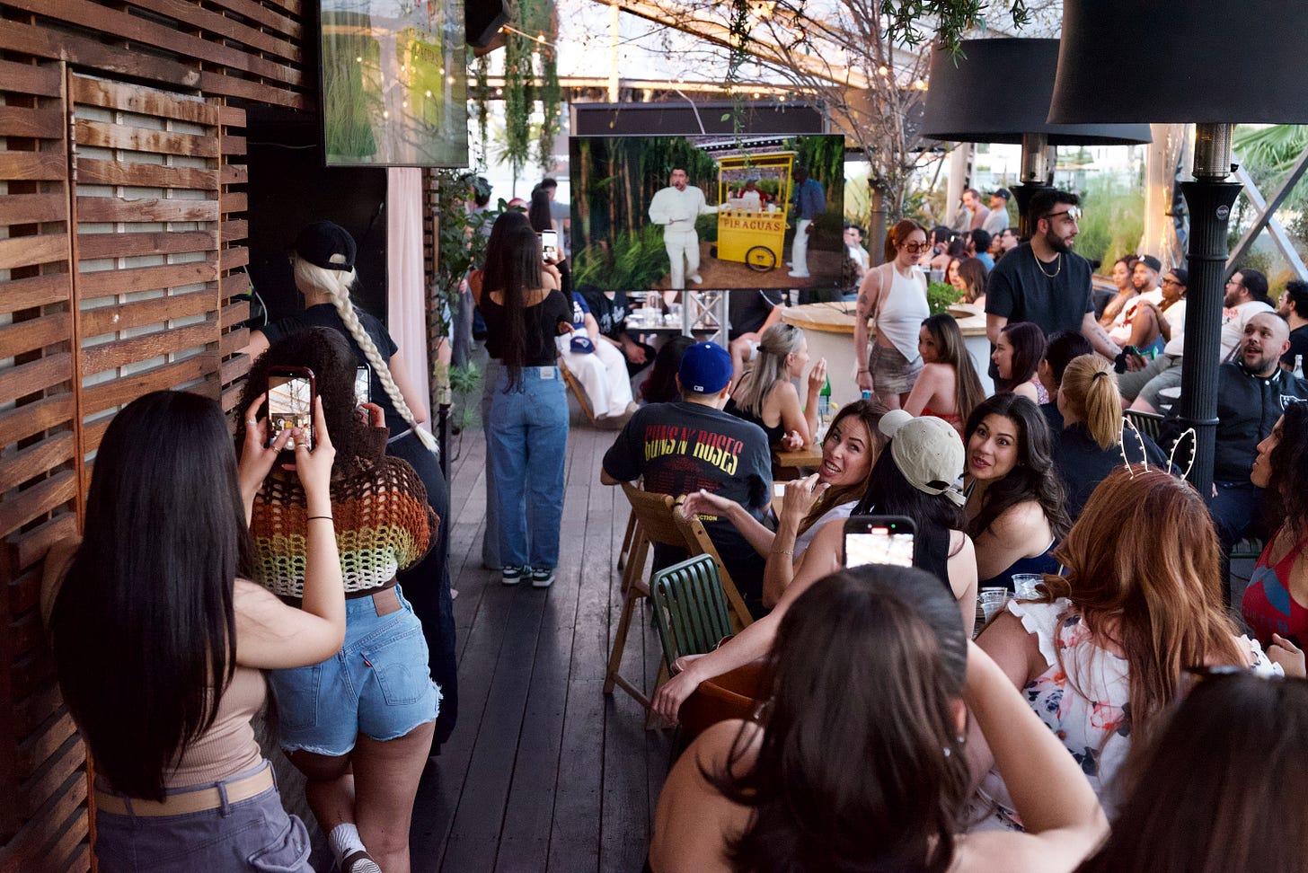 Wide rooftop scene at dusk, shot from behind the crowd as multiple guests lift their phones toward a television screen playing the Super Bowl; warm wood paneling lines the left frame while string lights and patio heaters create a layered glow, the performer bad bunny, visible only through the screen as attention converges forward.