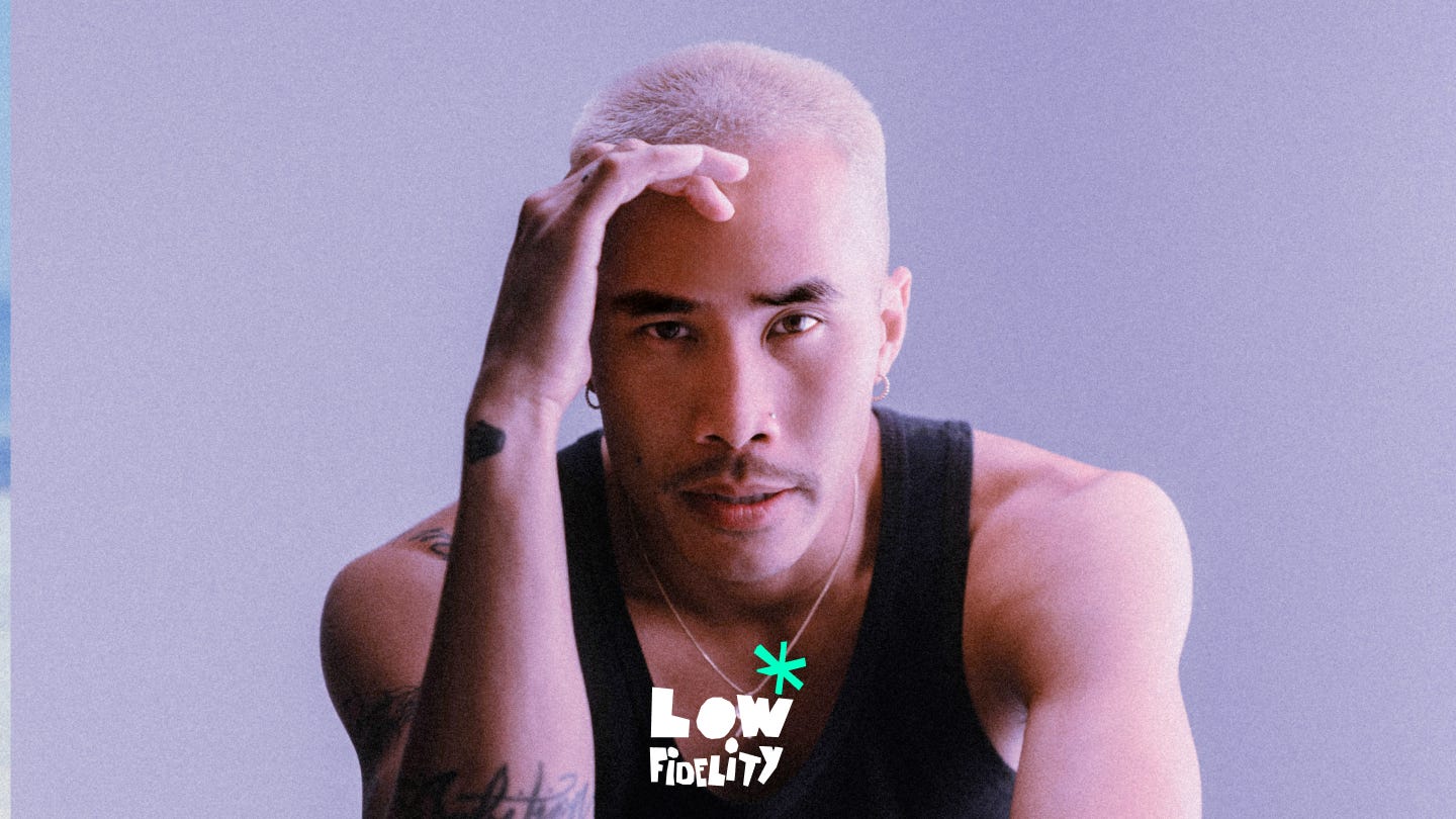 A portrait of a person with bleached buzz-cut hair leaning forward and looking directly at the camera against a soft purple background. They are wearing a black tank top and resting one hand on their head. The “Low Fidelity” logo appears centered near the bottom of the image in white text with a turquoise star-like symbol above it.