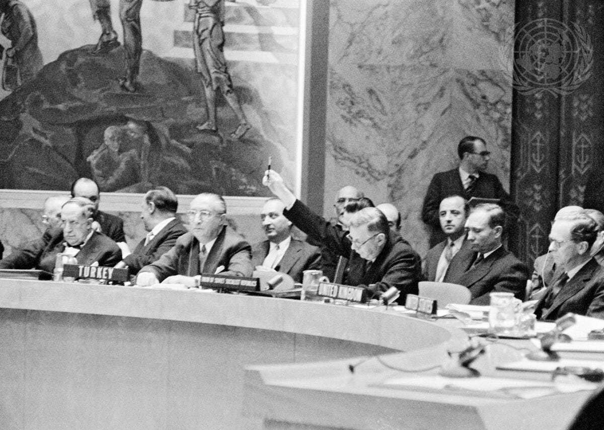 A delegate raises his hand at the Security Council table while other representatives sit alongside him, beneath a tall carved wall panel. A delegate raises his hand at the Security Council table while other representatives sit alongside him, beneath a tall carved wall panel.