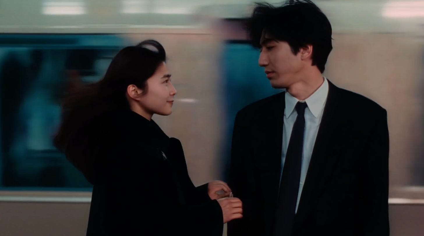 A woman and a man in a suit on a station platform as a train blurs past behind them, her hair blown by its wind