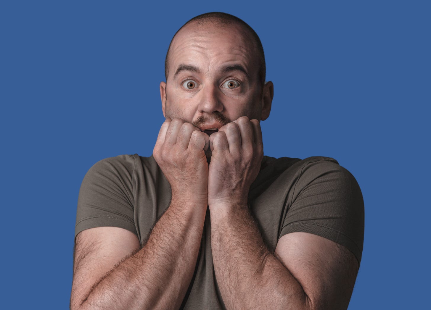 A bald dad wearing a t-shirt and his hands up to his mouth gasping with a scared or curious expression.