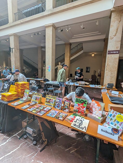 The first image shows a vibrant banner for the Unlock Book Fair in Munich, held on May 18-19, 2024, at the Abguss Museum. The banner is brightly colored with bold pink, yellow, and blue lettering, advertising the event as a feast of graffiti books and zines. The banner is hung above the entrance to a grand stone building with tall columns, as a person is seen checking their phone near the entrance. The second image captures a scene inside the book fair, where attendees browse through various graffiti books and zines at booths set up on long tables. In the foreground, a table prominently features Soi Books products, including zines, art books, and a selection of graffiti-related publications. The third image depicts two individuals at the book fair, one of whom is smiling and holding up a colorful graffiti art print by artist Tizer. The fourth image captures a lively moment in a traditional beer hall, where a group of people are enjoying drinks while a live band plays music in the background. One person is clapping along with the music, while others sit around the table chatting and drinking. The fifth image shows a classic Munich subway train arriving at a station, with its iconic blue and cream design. The train is captured just as it pulls into the platform, with a few passengers visible inside.