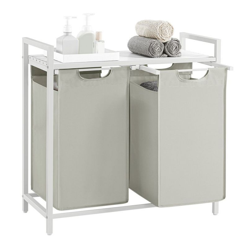 A dual-compartment beige colored laundry hamper with a white frame, topped with a tray holding rolled towels and pump bottles.