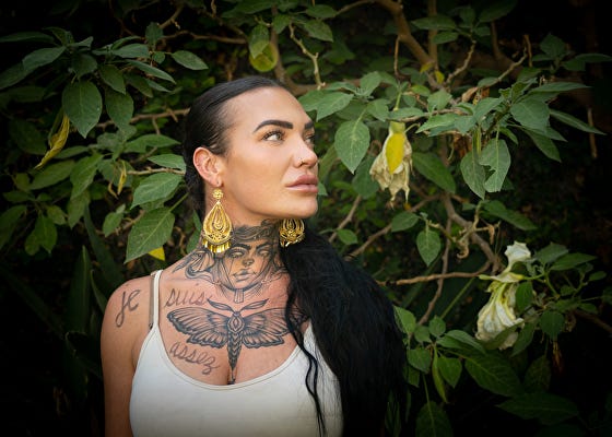 Woman in front of greenery looking to the left with 'je suis assez' written on her body in black ink
