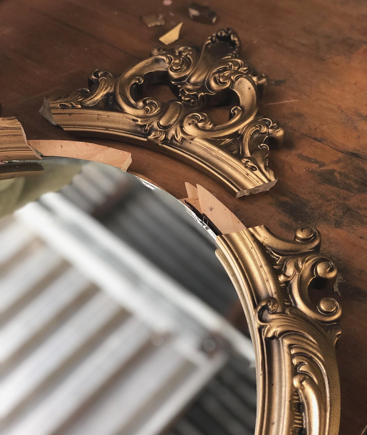 An oval mirror with an ornate antique gold frame that has a piece broken off, lying next to the frame