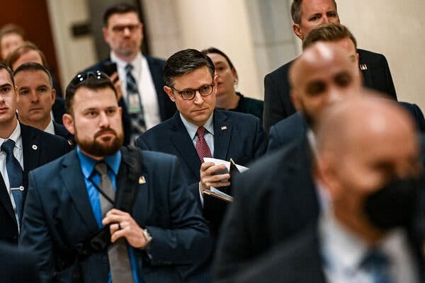 Speaker Mike Johnson walking in the middle of a group. He is wearing a dark suit, blue shirt and red tie. Speaker Mike Johnson walking in the middle of a group. He is wearing a dark suit, blue shirt and red tie.