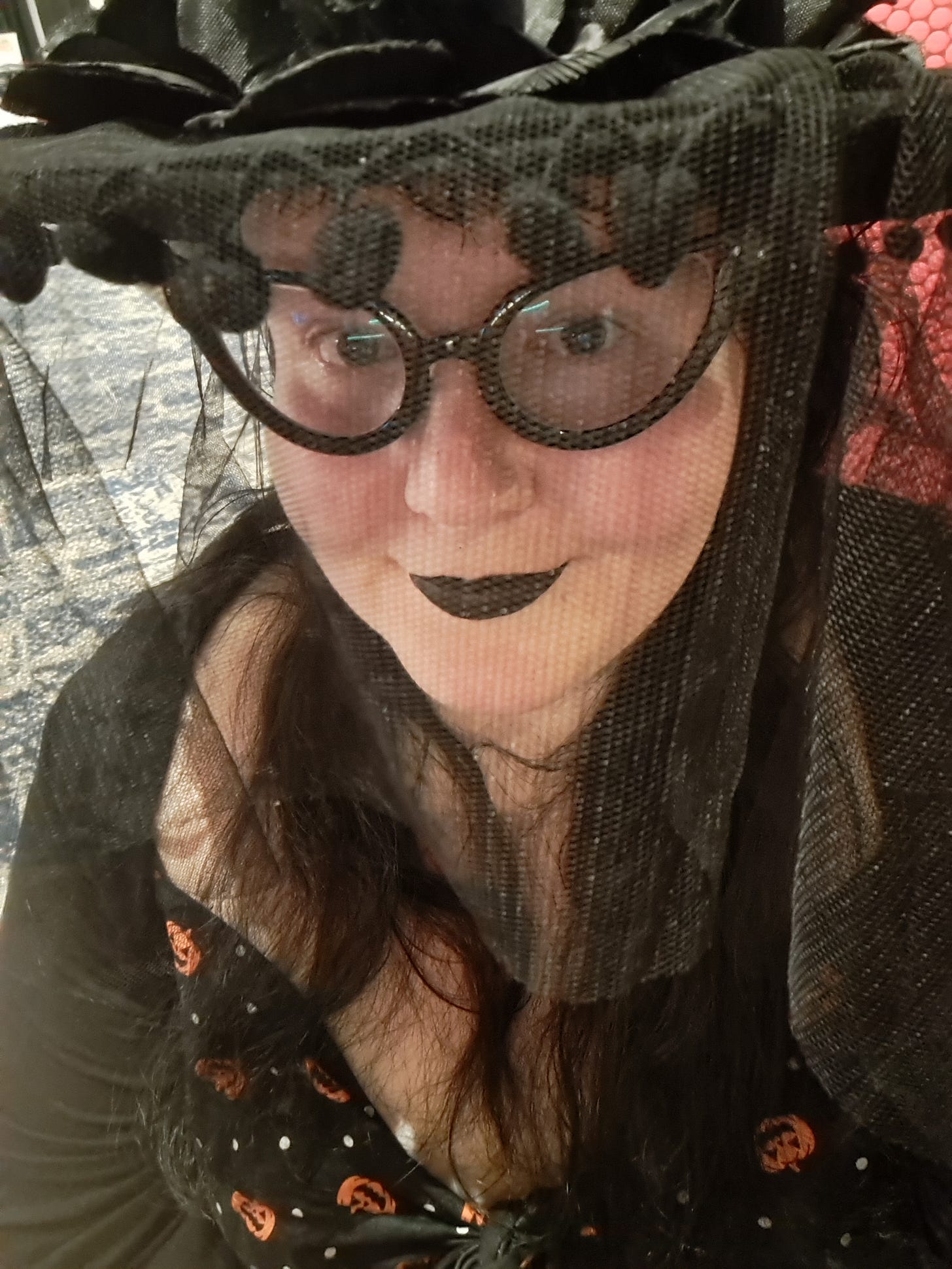 A selfie style picture of a woman in a black veiled hat, smiling while wearing black lipstick and a Halloween pumpkin dress.