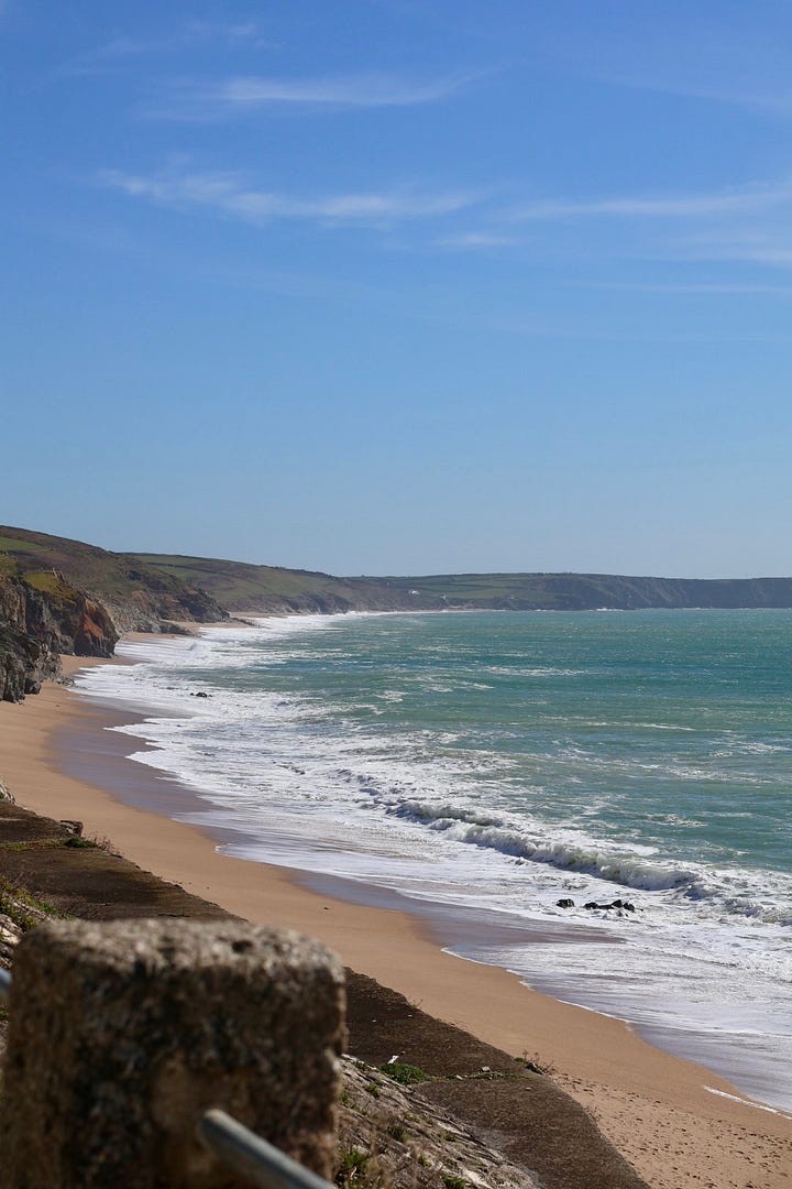 Coastal view of a sandy beach with waves rolling in beneath a clear blue sky, alongside a grassy cliffside overlooking the sea.