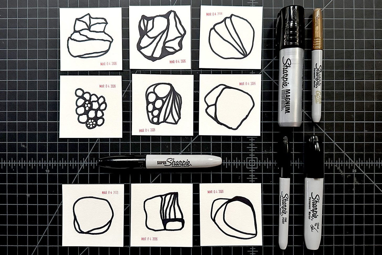 Nine small cards with abstract marker drawings arranged on a cutting mat, with Sharpie markers beside them. Nine small cards with abstract marker drawings arranged on a cutting mat, with Sharpie markers beside them.