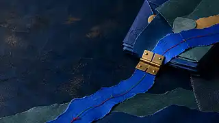 A folded cobalt paper path crosses a brass hinge, carrying visible thread from layered working material into a clear route.