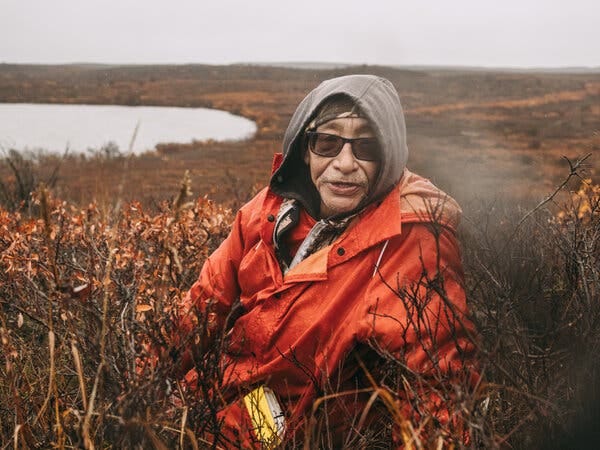 A man in an orange jacket, gray hoodie, and dark sunglasses stands amid dry, rust-colored brush.