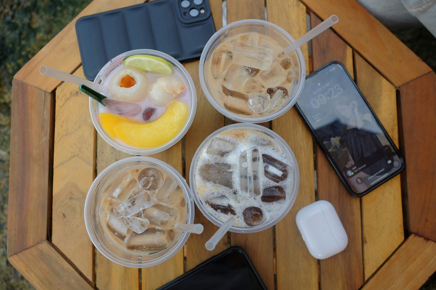 ​​A table topped with 4 plastic cups filled with drinks, two phones, and an AirPods case.
