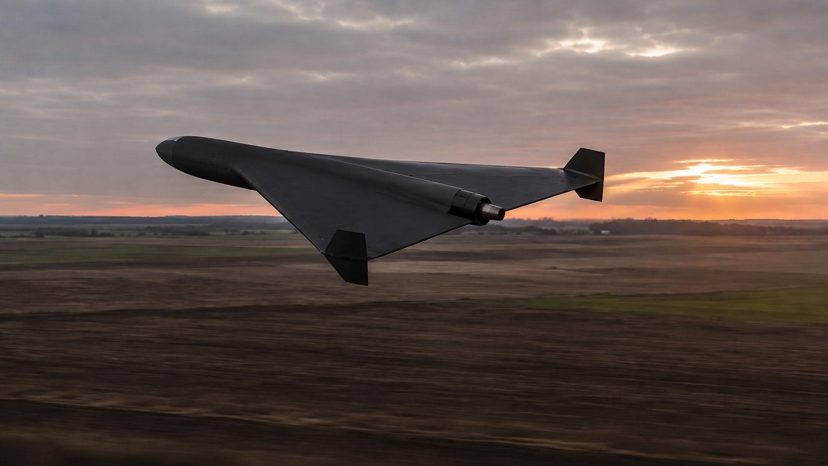 A black delta-wing drone with an external jet engine flies low over open farmland at dusk
