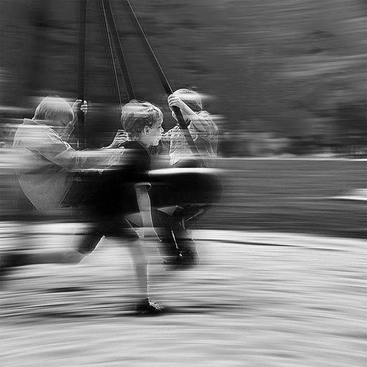 This may contain: black and white photograph of two people riding on a swing set with blurry trees in the background