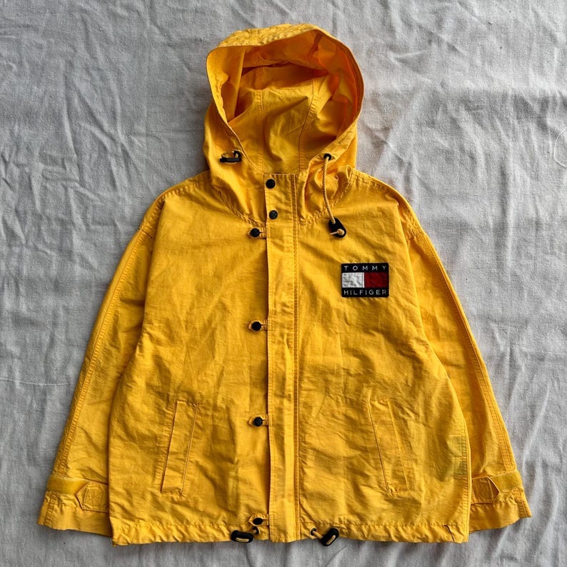 May include: A yellow hooded jacket with a Tommy Hilfiger logo on the left chest. The jacket has a drawstring at the bottom and a zipper closure.