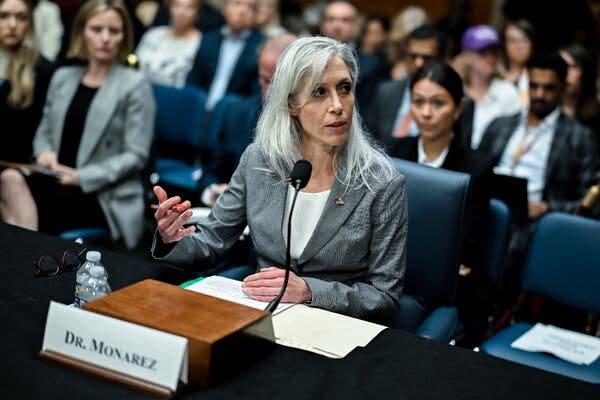A woman with gray slightly unkempt hair and a gray blazer sits behind a desk with a microphone and a placard that reads “Dr. Monarez.” A woman with gray slightly unkempt hair and a gray blazer sits behind a desk with a microphone and a placard that reads “Dr. Monarez.”