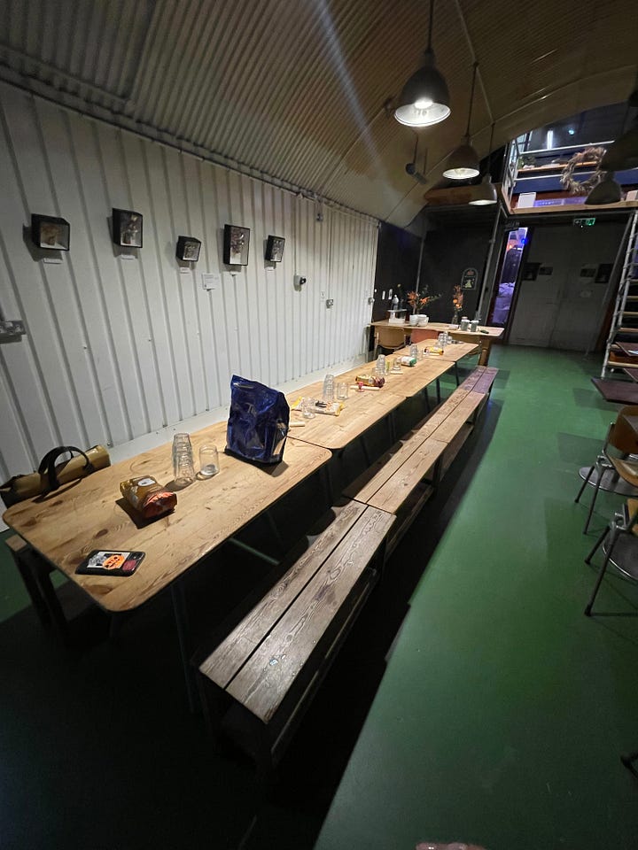 The first image is of a long wooden table with different bags of rice in an otherwise empty railroad arch. The second image is the same table with people walking around and examining eachothers rice artwork