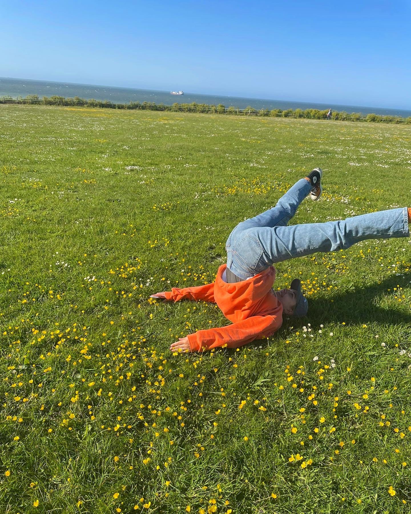 Tatum is on a patch of grass with the sea in the background. They are wearing an orange jumper and blue jeans and have their legs in the air 