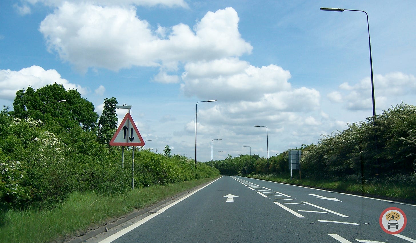 Photograph looking along a road from a driver's perspective. It is a single carriageway road with one lane each way, and painted arrows marking the two directions of travel. A warning sign on the left indicates two-way traffic. Photograph looking along a road from a driver's perspective. It is a single carriageway road with one lane each way, and painted arrows marking the two directions of travel. A warning sign on the left indicates two-way traffic.