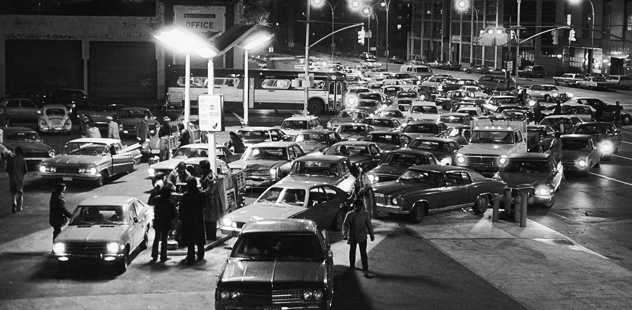 Black and white photograph from 1973 showing a crowded gas station at night during the OPEC oil embargo crisis. Dozens of cars are lined up bumper-to-bumper waiting to refuel, with drivers standing outside their vehicles. Street lights illuminate the chaotic scene with multiple lanes of traffic backed up. The image captures the panic and desperation of the 1973-1974 oil crisis when gas shortages paralyzed American motorists. While wildcatters rushed to drill oil wells during the boom years of 1976-1982, oilfield service companies like Schlumberger and Halliburton captured the real profits by selling essential services to all operators, eventually delivering 10x returns while most independent drillers went bankrupt when prices collapsed.