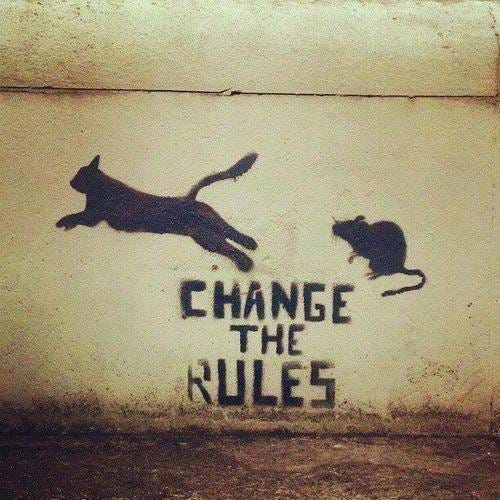 Black silhouette of a cat in mid-leap with extended tail and legs, pursuing a small mouse silhouette positioned to the right on a light beige wall surface. Bold black graffiti text above and below the figures spells Change the Rules in large capital letters with a graffiti style.