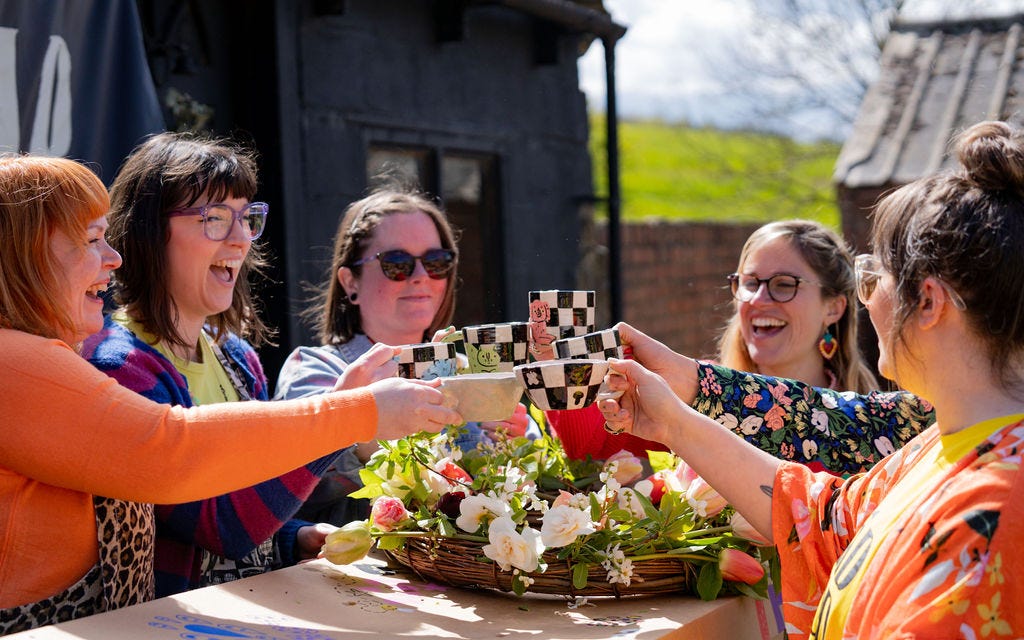 A group of friends raise a toast in chunky colourful mugs over a cardboard coffin and homemade wreath with flowers picked from the garden.