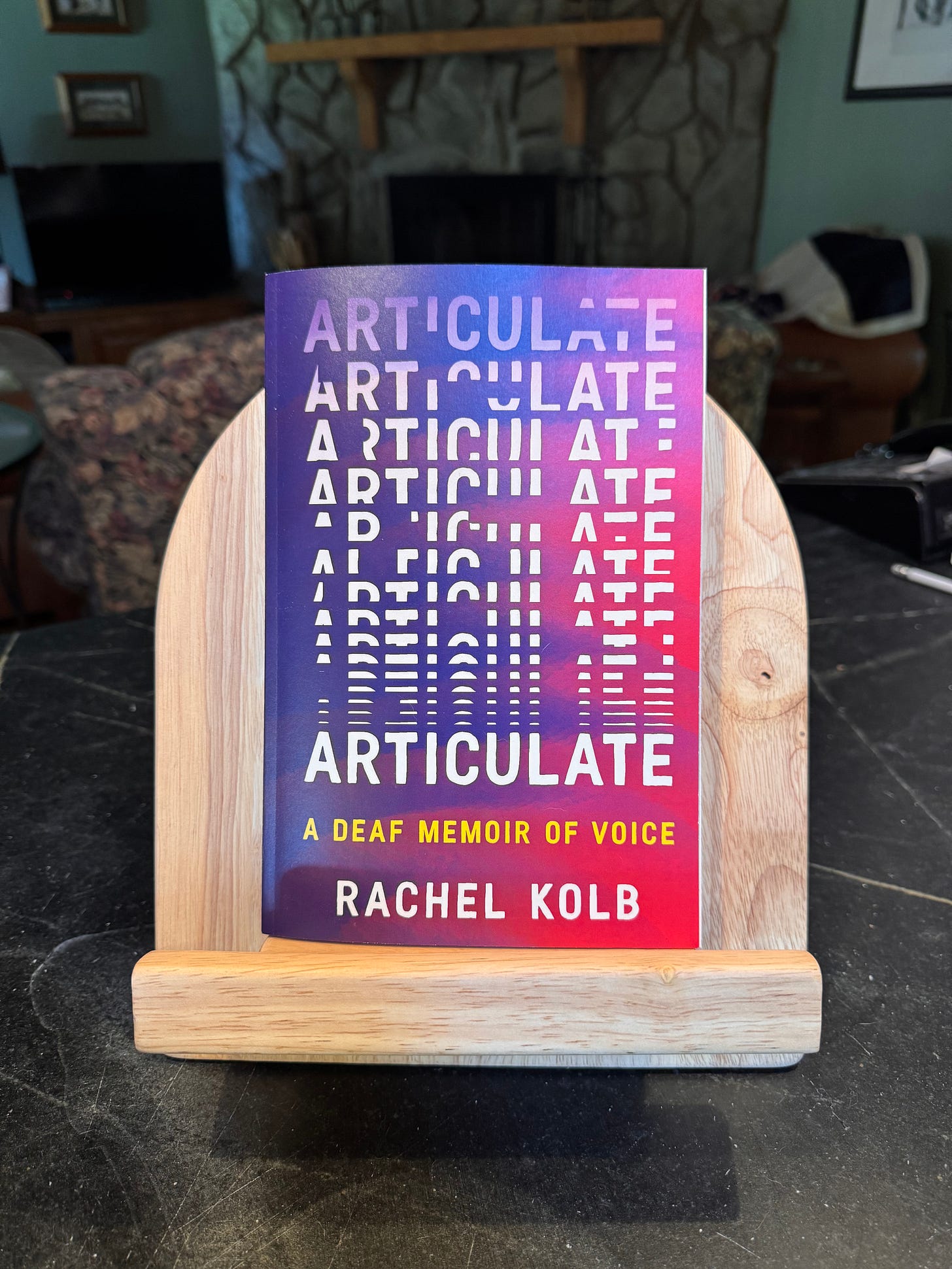 A galley copy of "ARTICULATE: A DEAF MEMOIR OF VOICE" sits on a wooden book pedestal on a counter. Cover features title letters assembling across the page, my name beneath, purple-and-pink swirls in the background.
