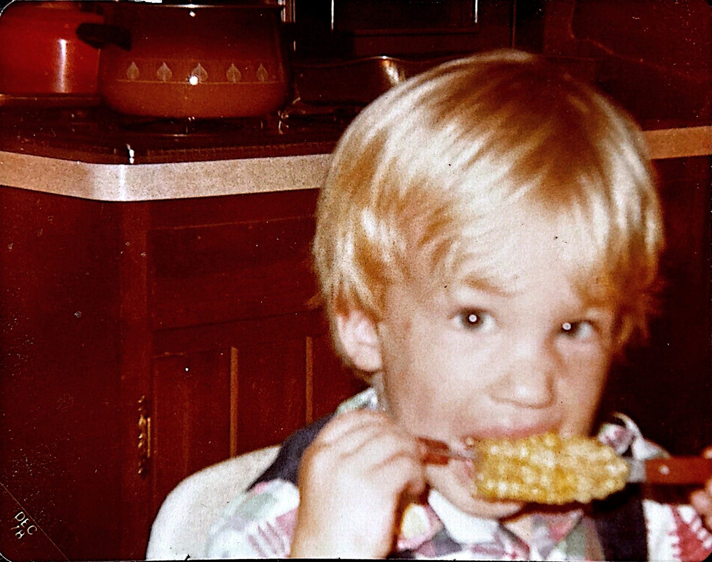 A child at a kitchen table, eating corn on the cob. The photo is slightly off-center, like a memory half-remembered.