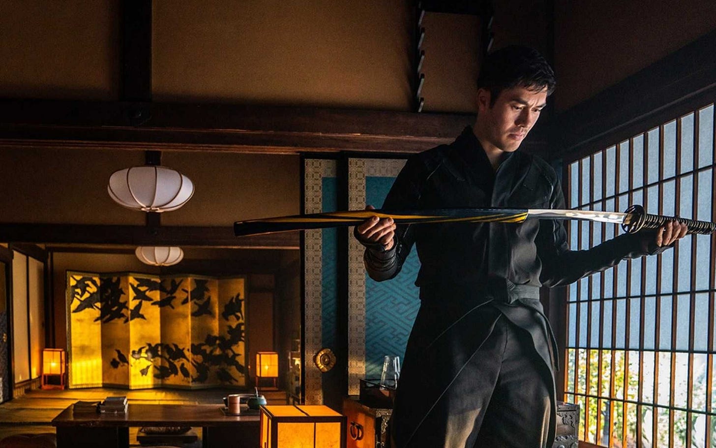 Henry Golding as Snake Eyes holding a sword halfway pulled out of its sheathe and looking down at it.