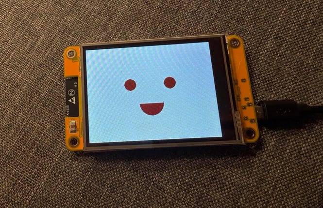 Photo of a yellow ESP32 touchscreen on a gray fabric surface displaying a white screen with a simple orange smiling face made of two eyes and a curved mouth.