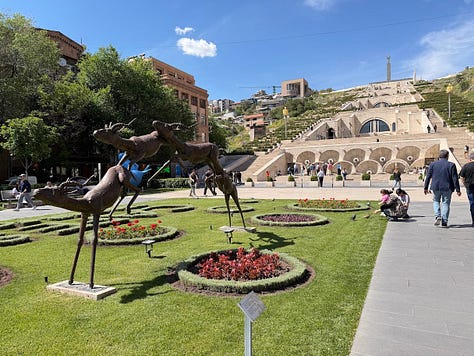 Scenes from around Armenia: bronze deer sculptures near the Cascade complex in Yerevan, a stone monastery nestled against forested mountains, a glass of amber brandy resting on an aging barrel, a hand holding a wine glass and folded lavash wrap at an outdoor food stand, bunches of grapes drying for raisining, and a colorful mural covering an apartment building facade in Yerevan