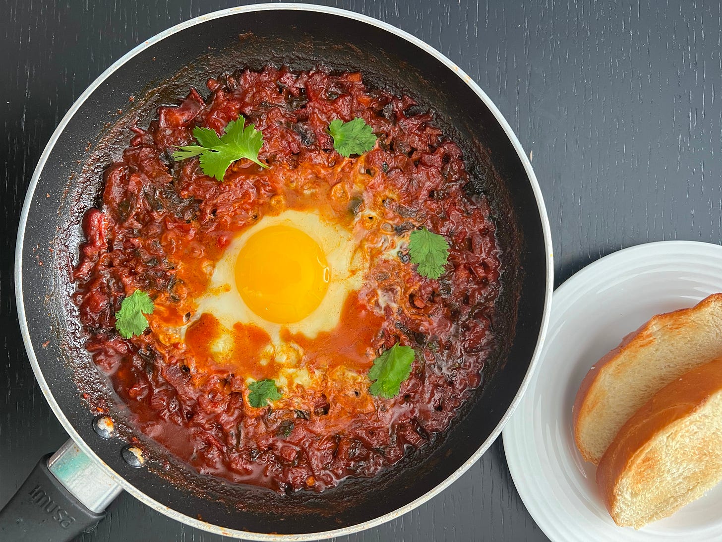 A skillet filled with shakshuka featuring a perfectly cooked egg in the center, topped with fresh cilantro leaves and served with slices of Challa bread on a white plate. This dish represents Liat Portal's ability to balance tradition with bold flavors on The Liat Show and Foodie Disorder.