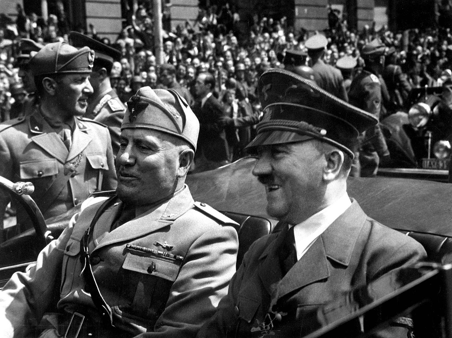 Benito Mussolini, in a military cap, and Adolf Hitler, in a peaked cap, sit side by side in an open car, smiling, with a crowd and uniformed men behind them.