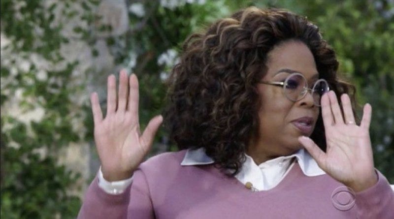 Oprah Winfrey wearing a lilac sweater and round black glasses, raising her hands in seeming disbelief and disgust