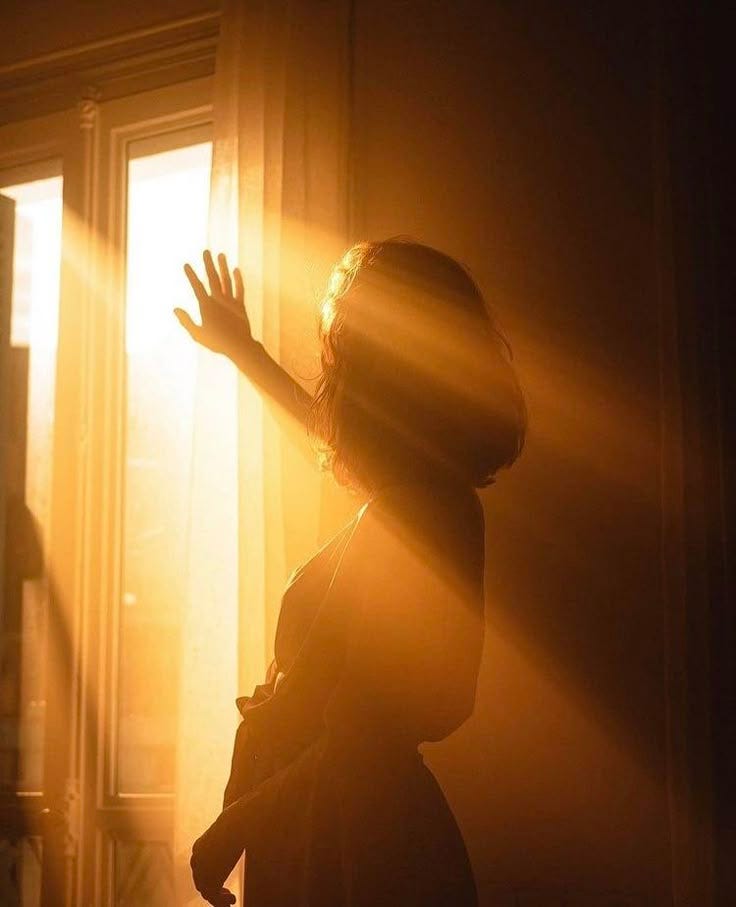 This may contain: a woman standing in front of a window with her hand up to the light coming through