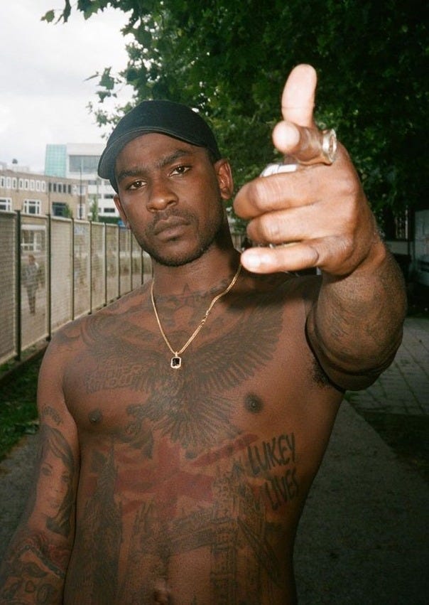 This may contain: a shirtless man with tattoos on his chest points at the camera while standing in front of a fence