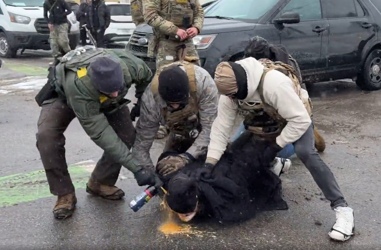 video screenshot of a male protester in a black winter coat and stocking cap being held face-down on the pavement by three ICE agents in camouflage and tactical vests while a fourth agent leans over to spray orange pepper spray directly into the protester's face. A small puddle of the orange liquid is left on the ground below the prone man's face. video screenshot of a male protester in a black winter coat and stocking cap being held face-down on the pavement by three ICE agents in camouflage and tactical vests while a fourth agent leans over to spray orange pepper spray directly into the protester's face. A small puddle of the orange liquid is left on the ground below the prone man's face.