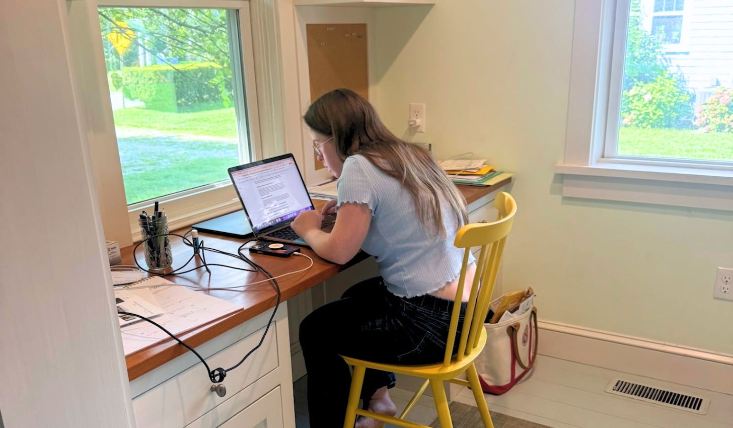Penny sits at a small desk in a bright nook, typing on her laptop, while windows and greenery outside are in the background